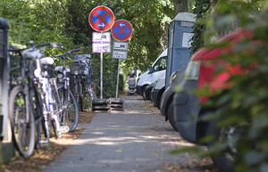 Fußweg: rechts stehen am Gartenzaun angelehnt Fahrräder, links sind Bäume und unbeschnittenem Grün, geparkte Autos und eine mobile Toilette.