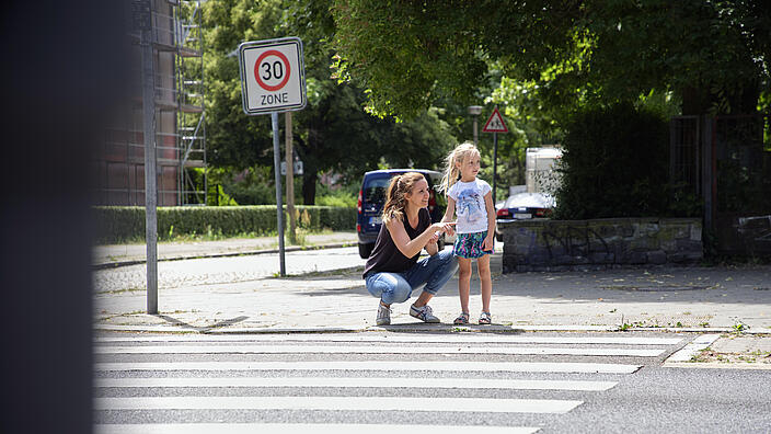 Eine Frau kniet neben einem Kind am Zebrastreifen. Sie zeigt mit der Hand nach rechts. Das Kind blickt ebenfalls nach rechts.