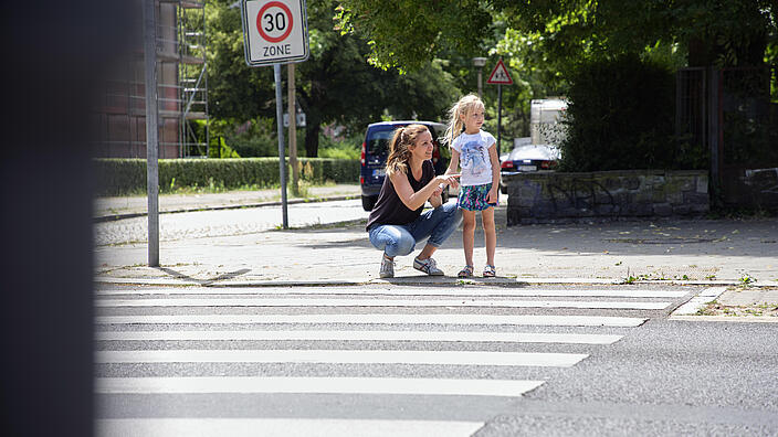 Eine Frau kniet neben einem Kind am Zebrastreifen. Sie zeigt mit der Hand nach rechts. Das Kind blickt ebenfalls nach rechts.