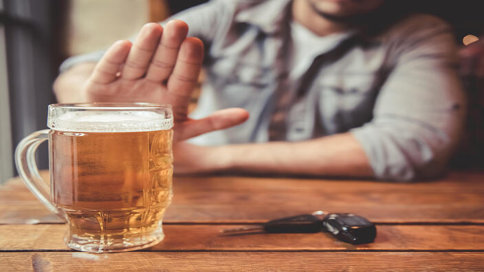 Eine Person sitzt an einem Holztisch und macht ein Handzeichen „Stopp“. Auf dem Tisch steht ein volles Bierglas und neben dem Glas liegt ein Autoschlüssel. 