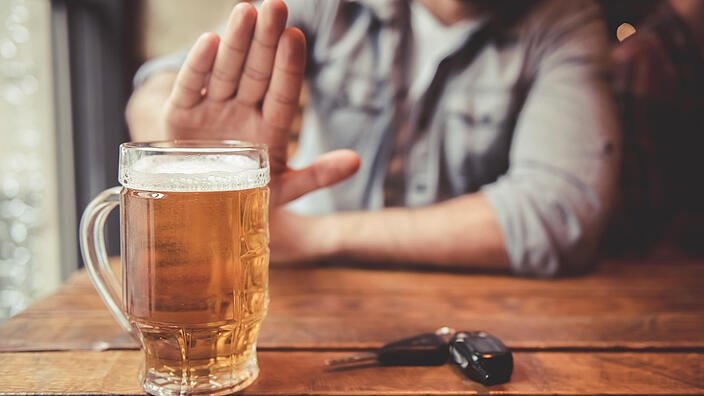 Eine Person sitzt an einem Holztisch und macht ein Handzeichen „Stopp“. Auf dem Tisch steht ein volles Bierglas und neben dem Glas liegt ein Autoschlüssel. 