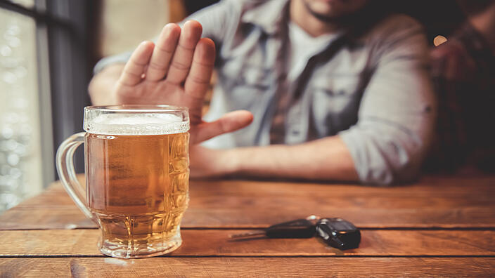 Eine Person sitzt an einem Holztisch und macht ein Handzeichen „Stopp“. Auf dem Tisch steht ein volles Bierglas und neben dem Glas liegt ein Autoschlüssel. 