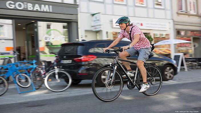 Ein Mann mit Helm, rosa Hemd und kurzen Hosen fährt mit einem Fahrrad entlang einer Straße mit Geschäften. Links im Bild ist ein Geschäft zu sehen, weitere abgestellte Fahrräder und ein schwarzes Auto.