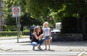 Eine Frau kniet neben einem Kind am Zebrastreifen. Sie zeigt mit der Hand nach rechts. Das Kind blickt ebenfalls nach rechts.
