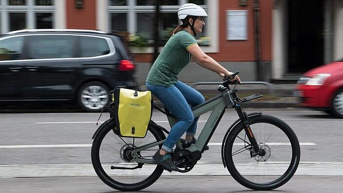 Eine Frau fährt auf einem Pedelec auf einem Radweg. Im Hintergrund sind zwei Parkende Autos zu sehen.