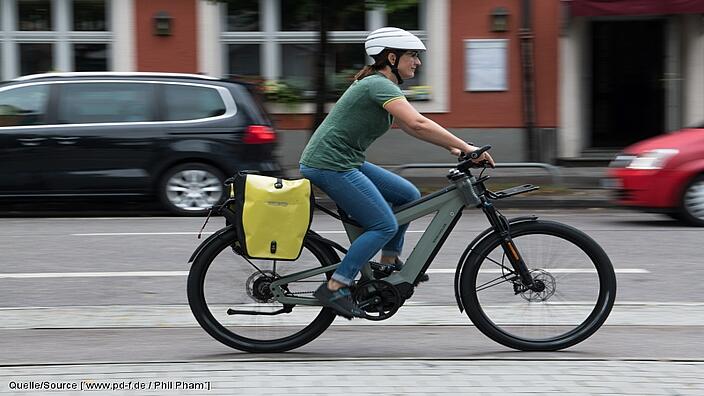 Eine Frau fährt auf einem Pedelec auf einem Radweg. Im Hintergrund sind zwei Parkende Autos zu sehen.