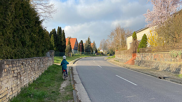 Landstraße in einem Ort. Kind fährt auf dem Trampelpfad neben der Straße, da es keinen Gehweg gibt.