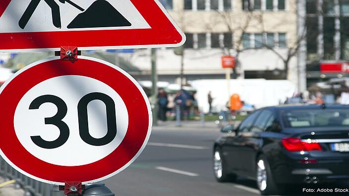 Am vorderen linken Bildrand ist ein Tempo 30-Schild. Dahinter fährt ein Auto entlang und Menschen stehen auf dem Gehweg. Der Hintergrund ist leicht verschwommen.