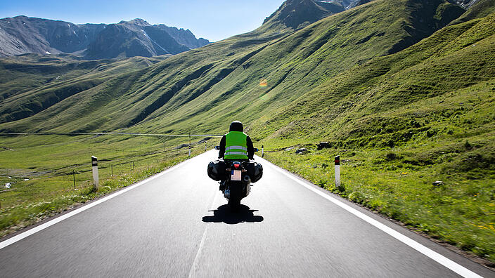 Ein Motorradfahrer fährt auf einer Landstraße durch die Berge. Die Sonne scheint.