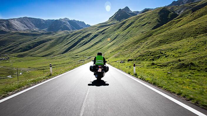 Ein Motorradfahrer fährt auf einer Landstraße durch die Berge. Die Sonne scheint.