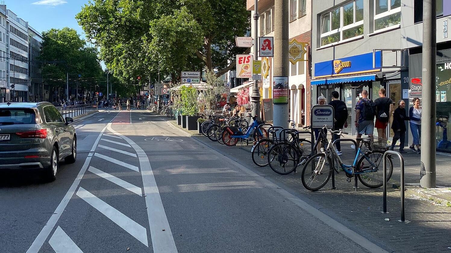 Sonderlösung im Querschnitt: ausgehend von der Knotenpunktausfahrt sind Taxistände, Fahrradparken und Liefern/Laden in Fahrbahnmitte angeordnet, vormalige Längsparkstände am Fahrbahnrand wurden mit Bügeln zum Abstellen von Fahrrädern ausgestattet oder für außengastronomische Nutzungen freigegeben; aus der Sperrfläche wird im weiteren Straßenver-lauf ein Schutzstreifen für geradeaus Radfahrende entwickelt (rechts im Hintergrund)