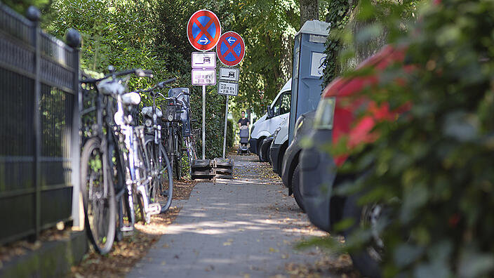 Fußweg: rechts stehen am Gartenzaun angelehnt Fahrräder, links sind Bäume und unbeschnittenem Grün, geparkte Autos und eine mobile Toilette.