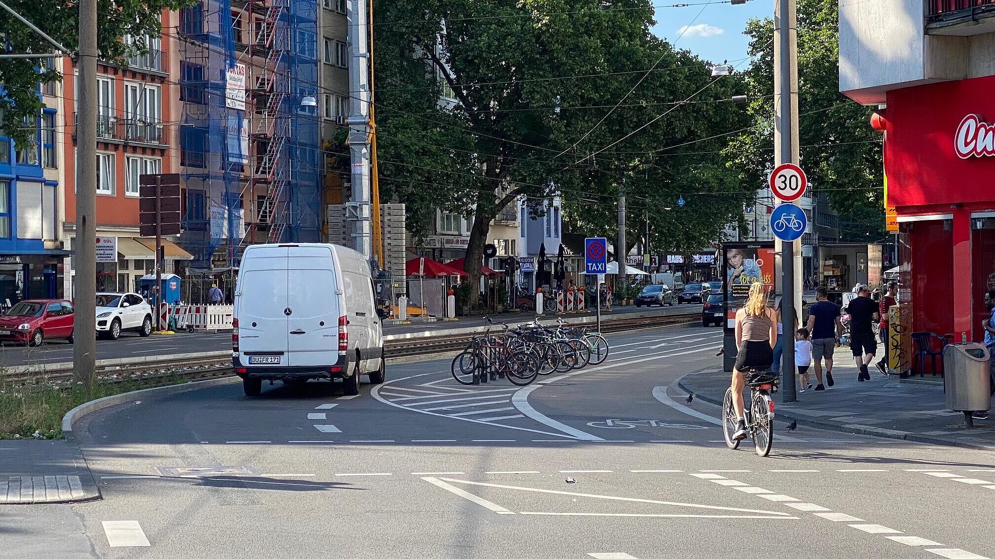 Sonderlösung im Querschnitt: ausgehend von der Knotenpunktausfahrt sind Taxistände, Fahrradparken und Liefern/Laden in Fahrbahnmitte angeordnet, vormalige Längsparkstände am Fahrbahnrand wurden mit Bügeln zum Abstellen von Fahrrädern ausgestattet oder für außengastronomische Nutzungen freigegeben