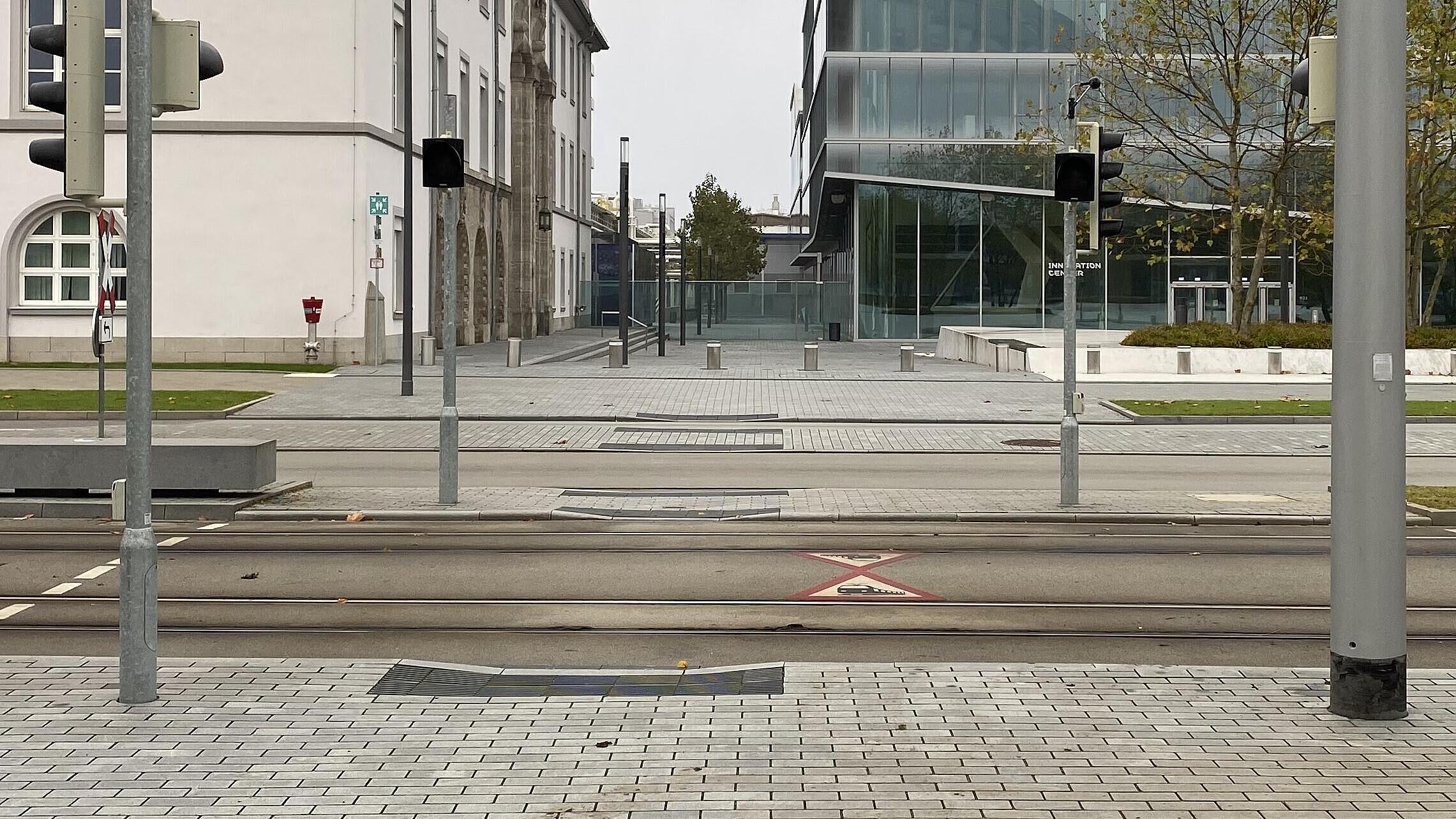 Querungsangebot: Dunkelampeln zeigen Gelbblinken bei Herannahen der Bahn, Rampen mit Bordabsenkung auf Null ermöglichen die barrierefreie Nutzung der Querungsstellen mit Rollstuhl und Rollator, Bodenindikatoren wurden zwecks besserer Erkennbarkeit für Sehbeeinträchtigte farblich konträr zum Oberflächenbelag gewählt – hier anthrazit auf hellgrau, weiß auf dunkelgrau