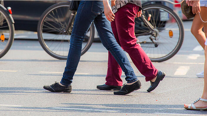 Drei Menschen überqueren eine Straße von rechts nach links. Man sieht nur ihre Beine und Füße.