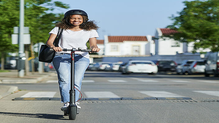 Eine Frau fährt mit einem E-Scooter auf der Straße. Dabei trägt sie einen Helm.