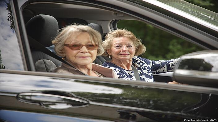 Zwei ältere Damen sitzen in einem schwarzen Auto. Eine von ihnen ist die Fahrerin des Pkws, die andere sitzt auf dem Beifahrersitz. Sie blicken in Richtung der Kamera, die ins Beifahrerfenster hinein fotografiert.