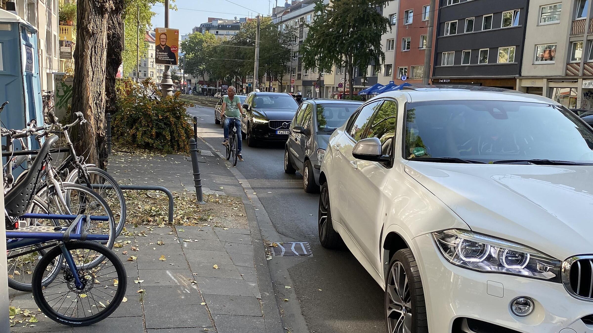 Bestandswahrung als absolute Ausnahme: Um den Kfz-Linksabbiegefahrstreifen und die vorhandenen Baum-reihe im angrenzenden Seitenraum beibehalten zu können, ist in einem kurzen Abschnitt des Karolingerrings der Schutzstreifen ausgesetzt; im Aufstellbereich vor der Lichtsignalanlage sind ein schmaler Streifen für links abbiegende und eine ebenso schmale Aufstellfläche für geradeaus Radfahrende, beide vorgezogen, markiert – eher symbolisch, da meist selbst von Pkw überfahren.