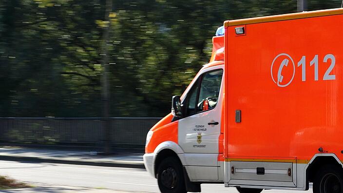 Ein Rettungswagen fährt mit Blaulicht über eine Straße. Die Bäume im Hintergrund sind aufgrund der Geschwindigkeit verwischt.