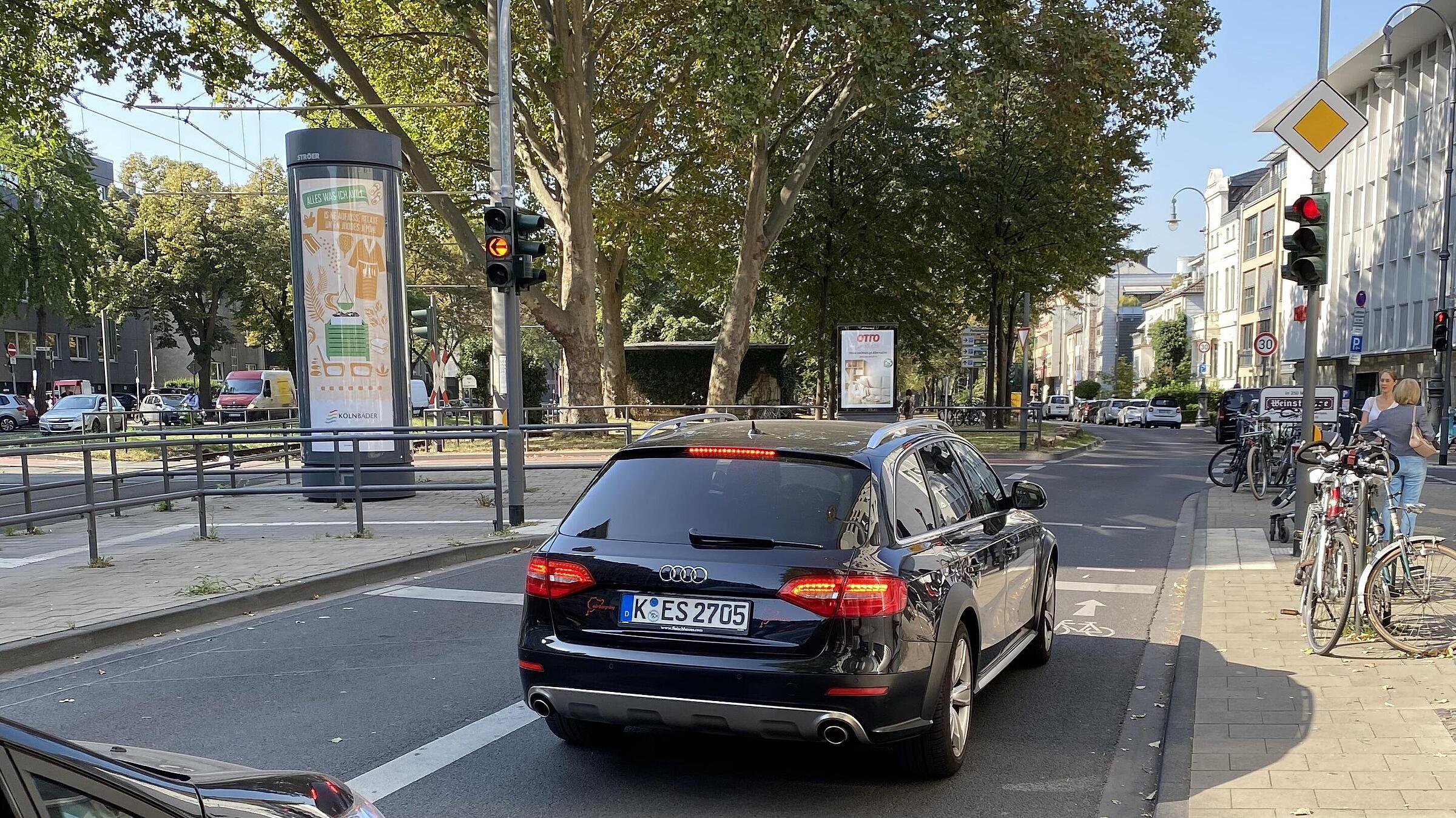 Bestandswahrung als absolute Ausnahme: Um den Kfz-Linksabbiegefahrstreifen und die vorhandenen Baum-reihe im angrenzenden Seitenraum beibehalten zu können, ist in einem kurzen Abschnitt des Karolingerrings der Schutzstreifen ausgesetzt; im Aufstellbereich vor der Lichtsignalanlage sind ein schmaler Streifen für links abbiegende und eine ebenso schmale Aufstellfläche für geradeaus Radfahrende, beide vorgezogen, markiert – eher symbolisch, da meist selbst von Pkw überfahren.