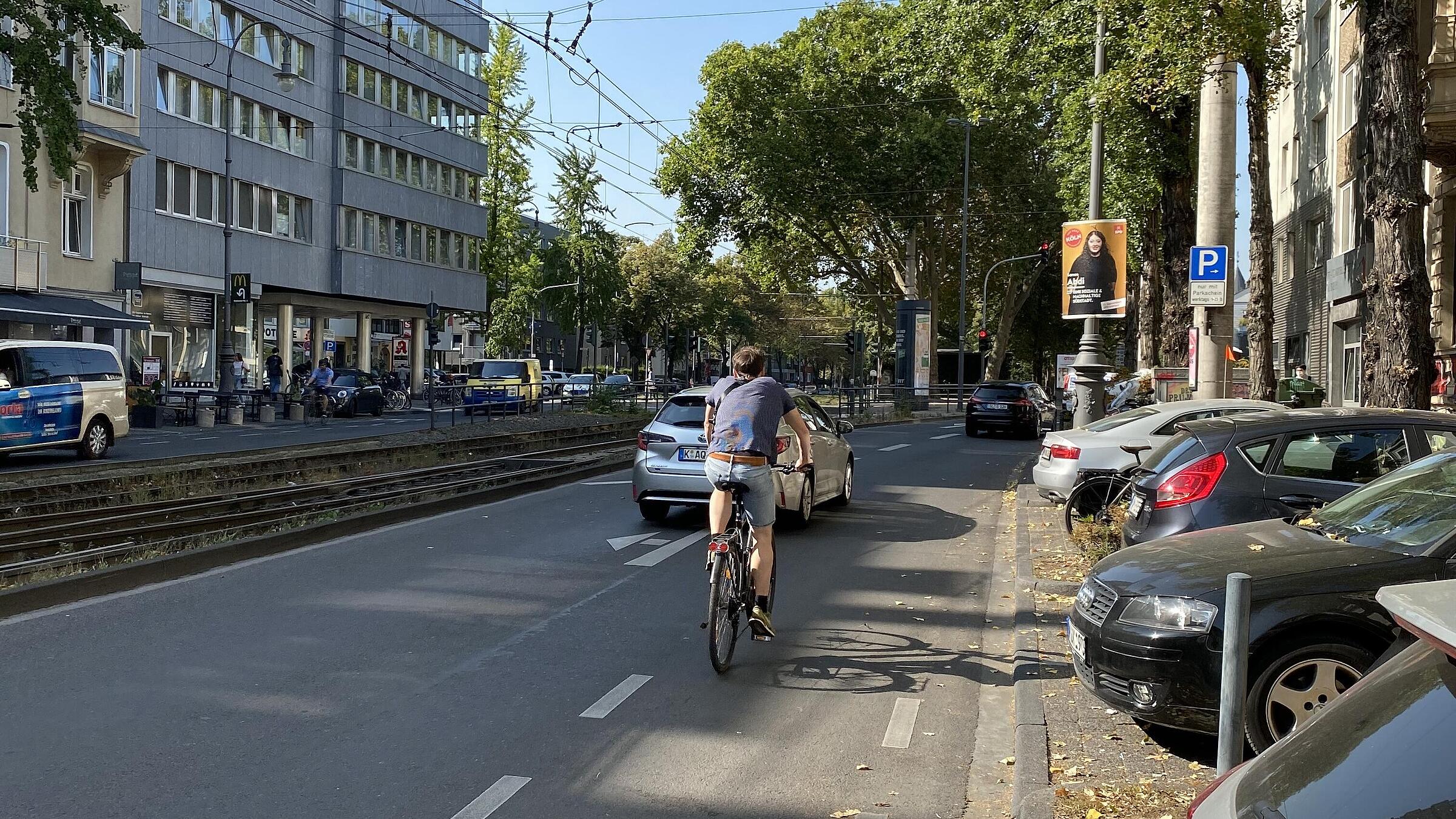 Bestandswahrung als absolute Ausnahme: Um den Kfz-Linksabbiegefahrstreifen und die vorhandenen Baumreihe im angrenzenden Seitenraum beibehalten zu können, ist in einem kurzen Abschnitt des Karolingerrings der Schutzstreifen ausgesetzt; im Aufstellbereich vor der Lichtsignalanlage sind ein schmaler Streifen für links abbiegende und eine ebenso schmale Aufstellfläche für geradeaus Radfahrende, beide vorgezogen, markiert – eher symbolisch, da meist selbst von Pkw überfahren.