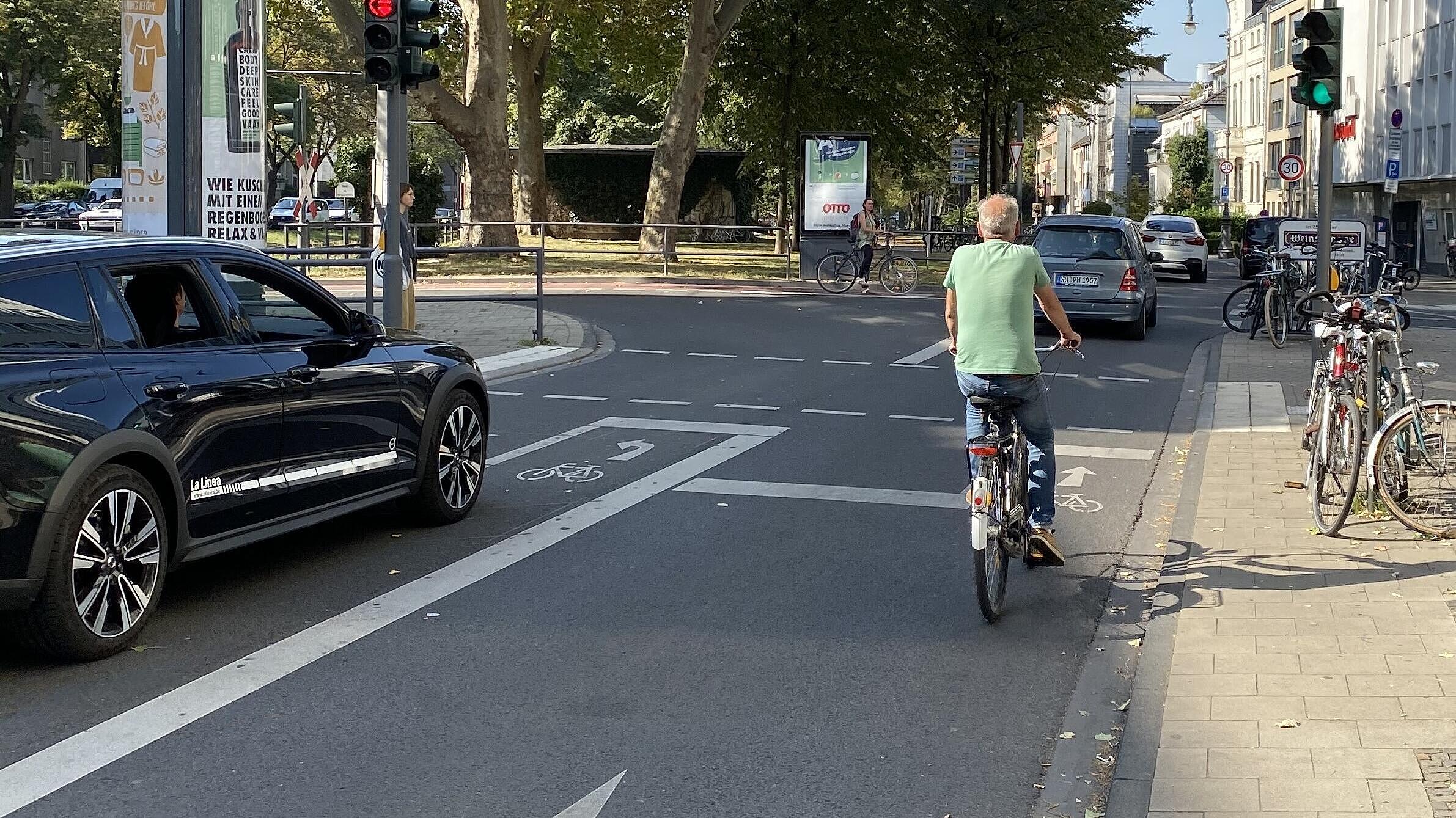 Bestandswahrung als absolute Ausnahme: Um den Kfz-Linksabbiegefahrstreifen und die vorhandenen Baum-reihe im angrenzenden Seitenraum beibehalten zu können, ist in einem kurzen Abschnitt des Karolingerrings der Schutzstreifen ausgesetzt; im Aufstellbereich vor der Lichtsignalanlage sind ein schmaler Streifen für links abbiegende und eine ebenso schmale Aufstellfläche für geradeaus Radfahrende, beide vorgezogen, markiert – eher symbolisch, da meist selbst von Pkw überfahren.