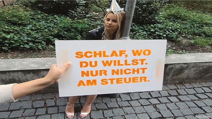 Eine Frau lehnt mit geschlossenen Augen an einen Laternenmast. Sie trägt einen Partyhut. Vor ihr steht ein Schild mit der Aufschrift "Schlaf, wo du willst. Nur nicht am Steuer".