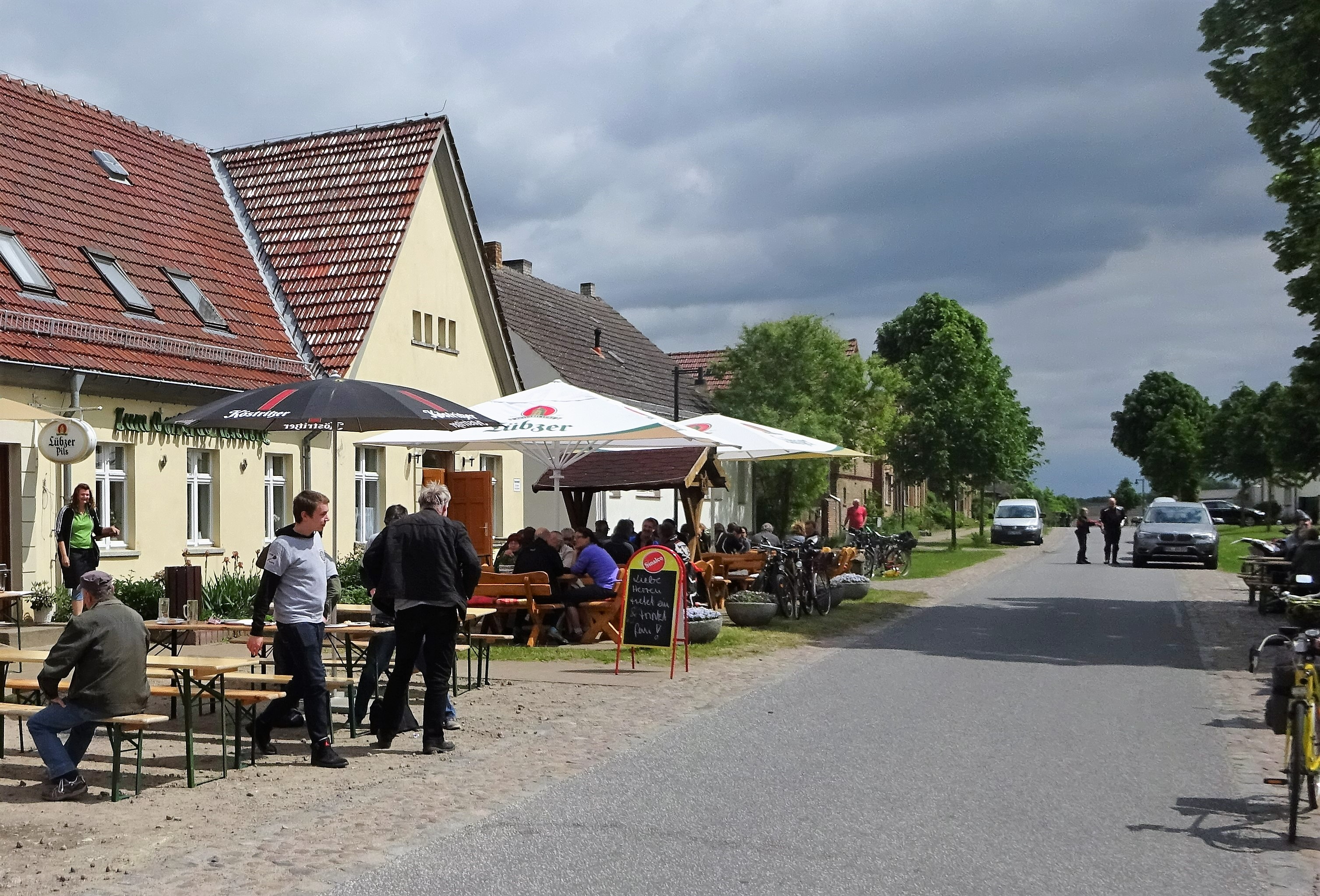 Bild 19: Hausvorbereich und Ausweichstelle: Außengastronomie am Dorfkrug mit Parkmöglichkeit für Gäste auf der gegenüber liegenden Pflasterfläche, die ursprünglich als Ausweichfläche bei der Begegnung von breiteren Fahrzeugen geplant war